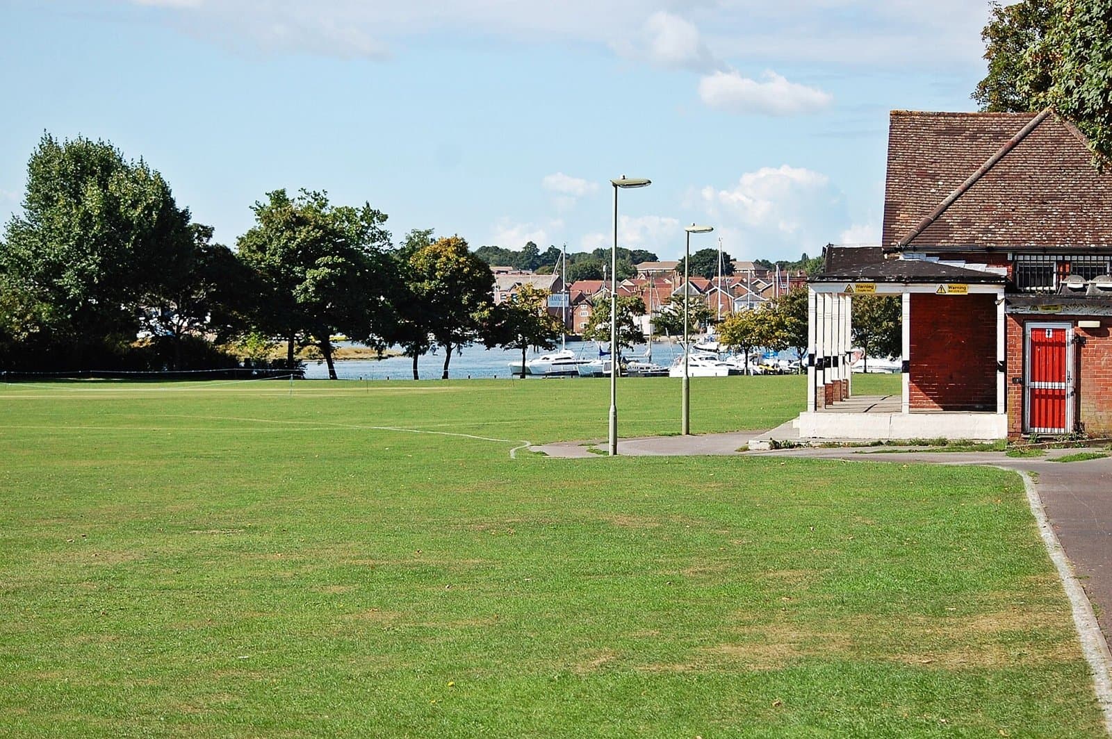 Open grass at Bath Lane Recreation Ground, Fareham, with a pavilion and moored boats on the creek behind