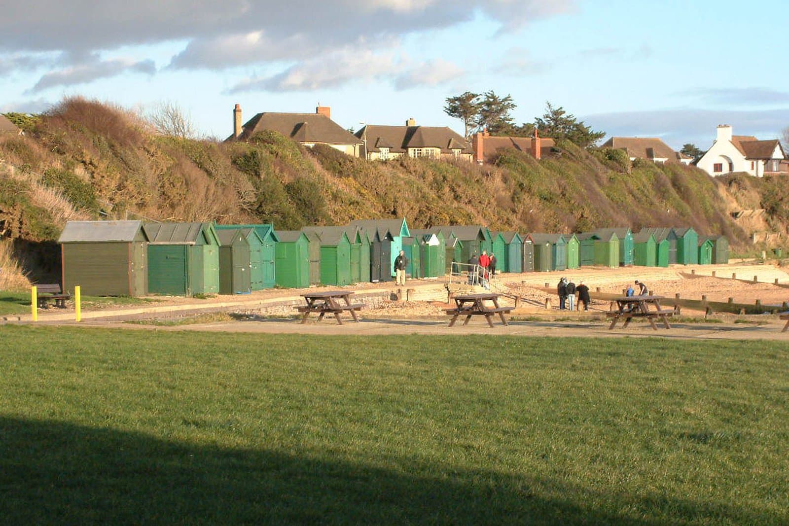 Row of green beach huts below the cliff at Hill Head, Hampshire, with picnic tables and lawn in the foreground